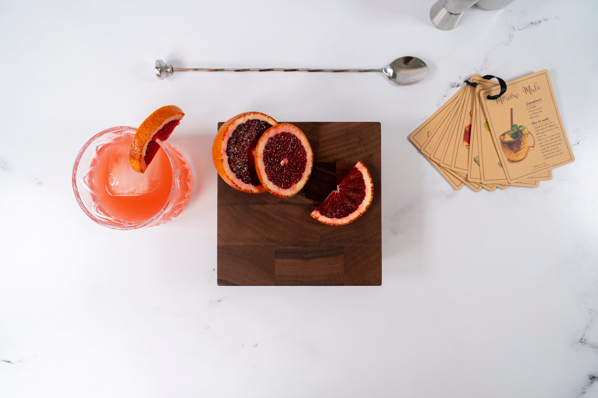 close-up of a square wooden cutting board by Square Root Studio, showcasing several vibrant slices of blood orange. The board rests on a clean white surface, highlighting the natural grain of the walnut and the bright citrus fruit. 6x6 end grain cutting board for cocktail enthusiast, cocktail and mocktail recipe cards, cocktail stirring utensils
