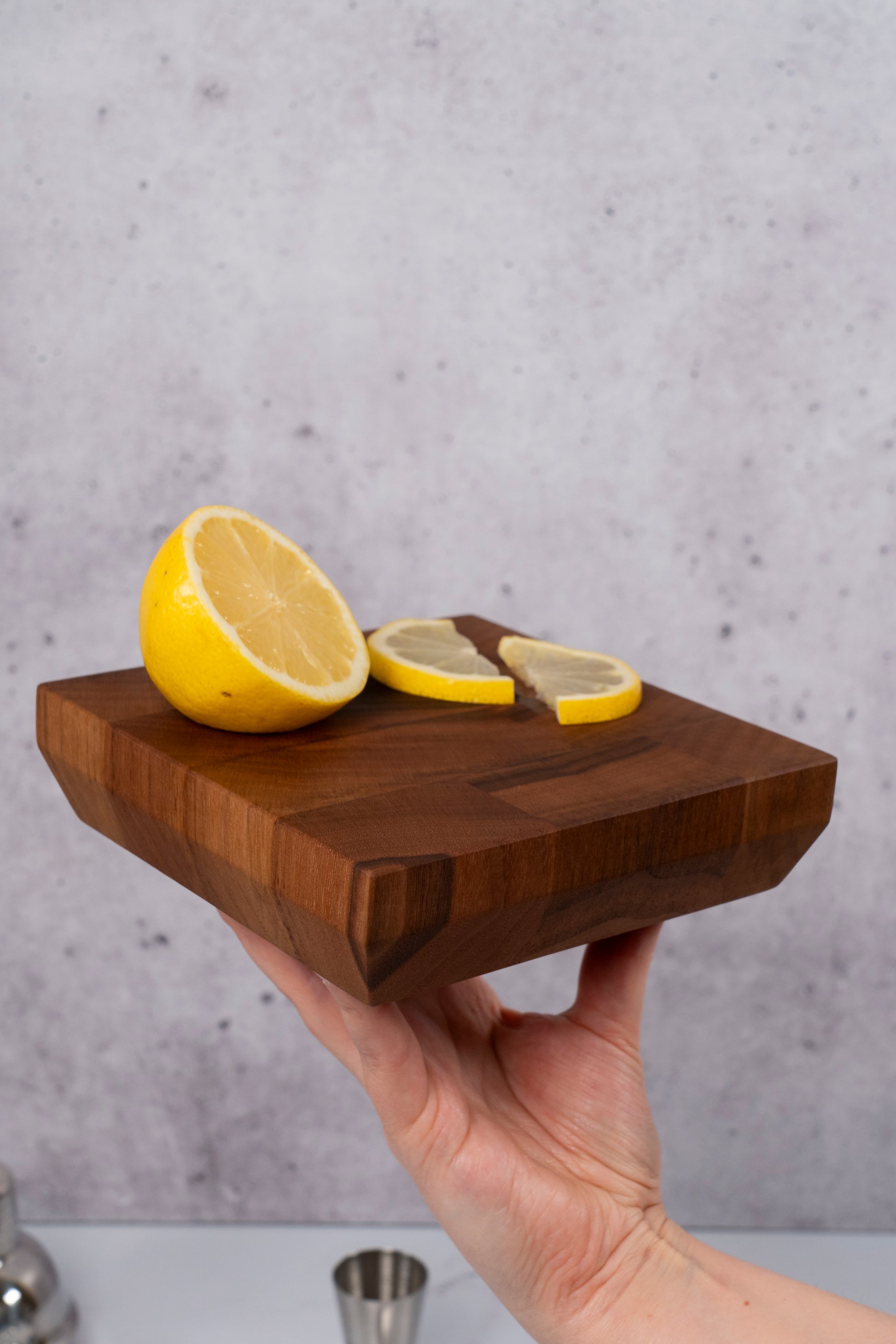 Small end grain cutting board made out of black american walnut with tapered edges, fresh cut lemon slices sitting on top, help in a single hand for display. Home bar and restaurant grade mini end-grain block ideal for bartender gift and cocktail enthusiasts made by Square Root Studio, made in USA. The tapered edges showcase attention to detail, and high quality craftsmanship