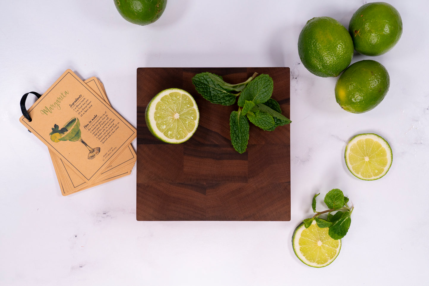 Cocktail of mixed drink bar board, walnut end grain board 6 by 6 inches and 1.5 inches thick placed on marble counter top along cocktail recipes, has cut limes and a mint cutting on top. Home bar mixology tool made by Square Root Studio, ideal for parties at home and cocktail preparations.