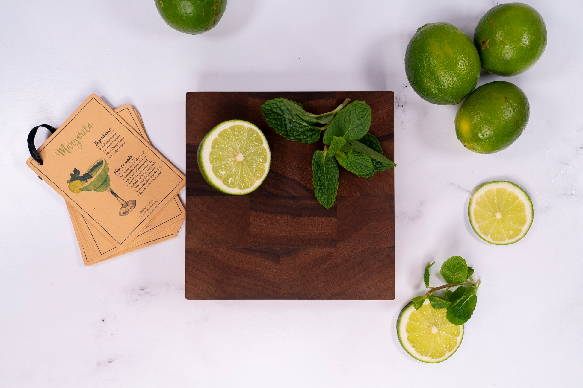 Cocktail of mixed drink bar board, walnut end grain board 6 by 6 inches and 1.5 inches thick placed on marble counter top along cocktail recipes, has cut limes and a mint cutting on top. Home bar mixology tool made by Square Root Studio, ideal for parties at home and cocktail preparations.