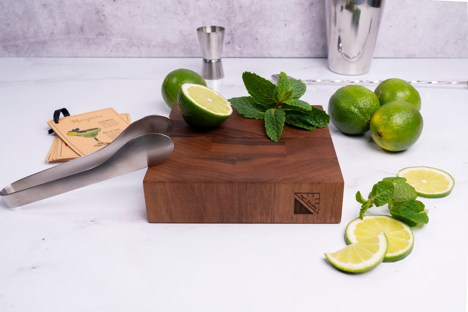 A rustic wooden Square Root Studio cutting board displays fresh cut lime slices and fresh mint leaves, possibly in preparation for a margarita drink. The setting suggests a stylish and inviting atmosphere for enjoying a handcrafted drink. Mini-chopper for cocktail and mocktail enthusiast, premium bar accessory