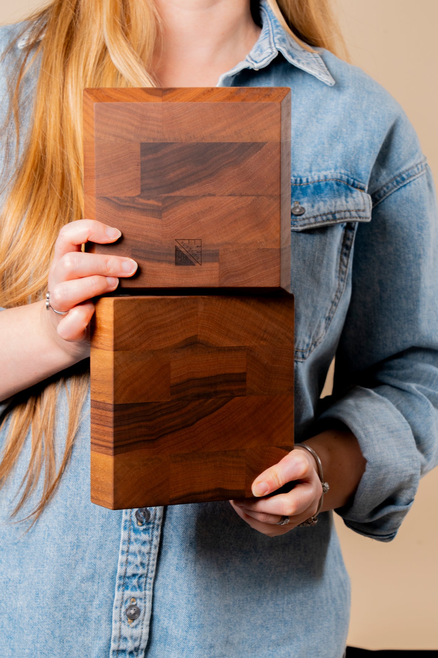 Two walnut end grain boards with sqrtStudio logo engraved on the bottom held by a young lady dressed in a relaxed blue jeans shirt. She wears silver jewelry, representing a sophisticated look. Two options of 6x6 walnut bar blocks with tapered or straight edges created as mixology accessory for bartender and cocktail enthusiasts.Premium bar block made in USA