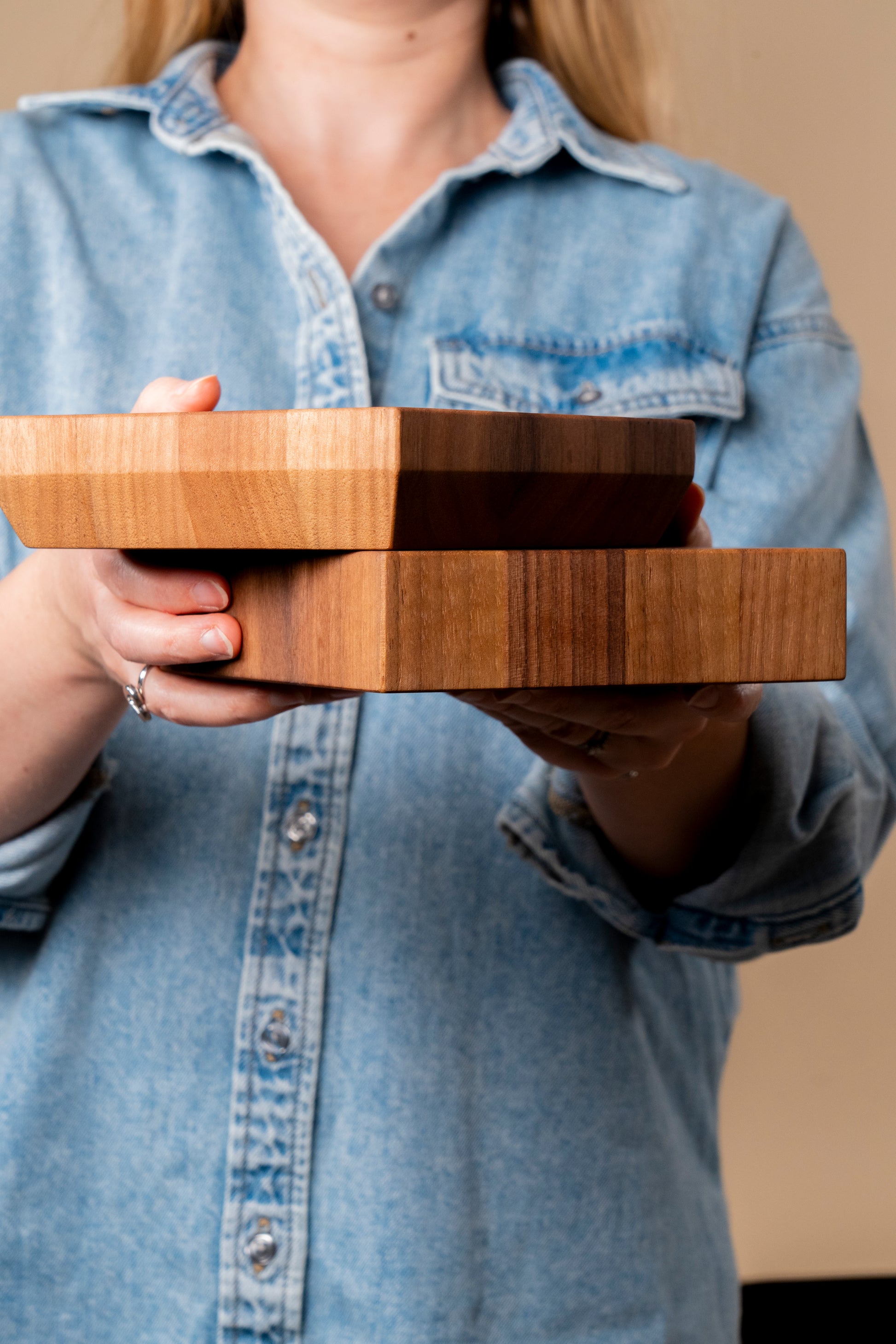 A young women dressed in blue jeans shirt holding two walnut end grain boards in her hands, one on top of each other. The end grain boards have straight and tapered edges, showcasing great craftsmanship and attention to detail. The boards are made out of high quality walnut, 100% made in USA by Square Root Studio, a premium brand for kitchen and home accessories.