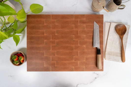 Premium extra large 20x15 maple end grain butcher block with rubber feet, Japanese steel knife with wooden handle, wooden spoon, bowl with fresh strawberries on a marble kitchen countertop. two brown ceramic bowls next to end grain cutting board. End grain cutting board sitting next to a green plant, hard maple cutting board ideal gift for cooking experts, wooden gift for 5th anniversary, high-quality and durable cutting board, sustainable BPA-free cutting