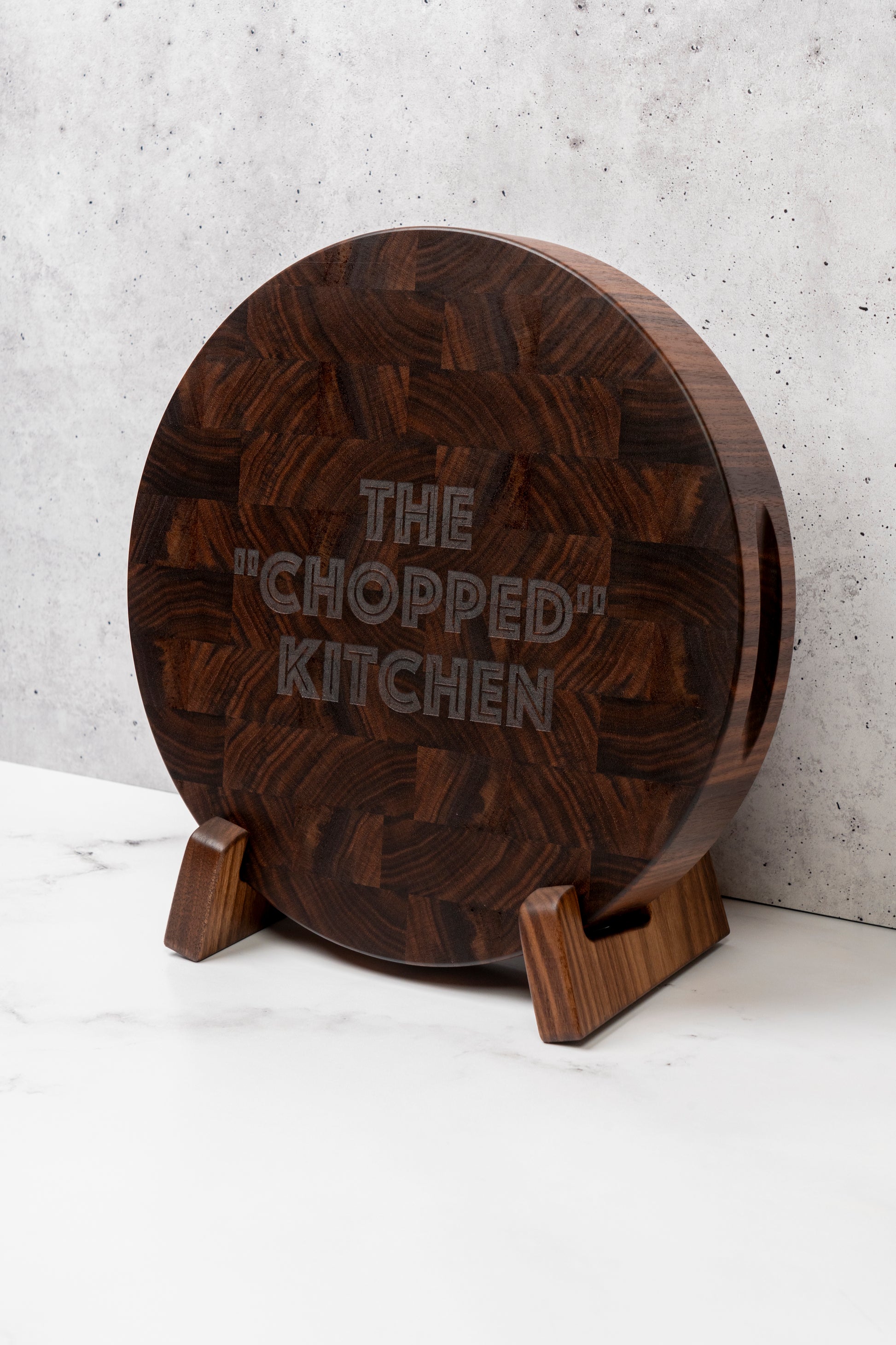 round solid wood butcher block, custom made end grain cutting board with the text "the chopped kitchen" message engraved on the front. end grain cutting board sitting vertically in a walnut cutting board stand with variable width, custom made by Square Root Studio in the USA, round end grain cutting board with handles